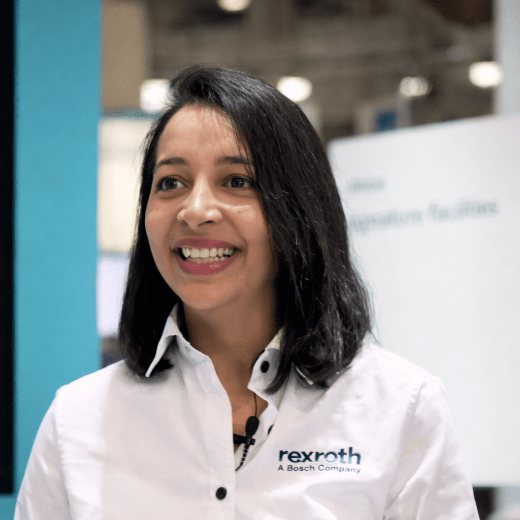 A woman with shoulder-length dark hair smiles in a white “rexroth A Bosch Company” shirt, indoors near Trackunit IrisX tech.