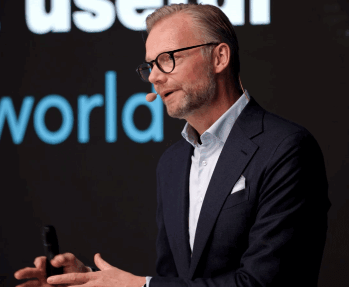 A man in glasses and a suit gestures while speaking on stage at the Trackunit Next event, related to construction IoT.
