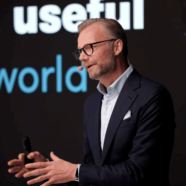 A man in glasses and a suit gestures while speaking on stage at the Trackunit Next event, related to construction IoT.