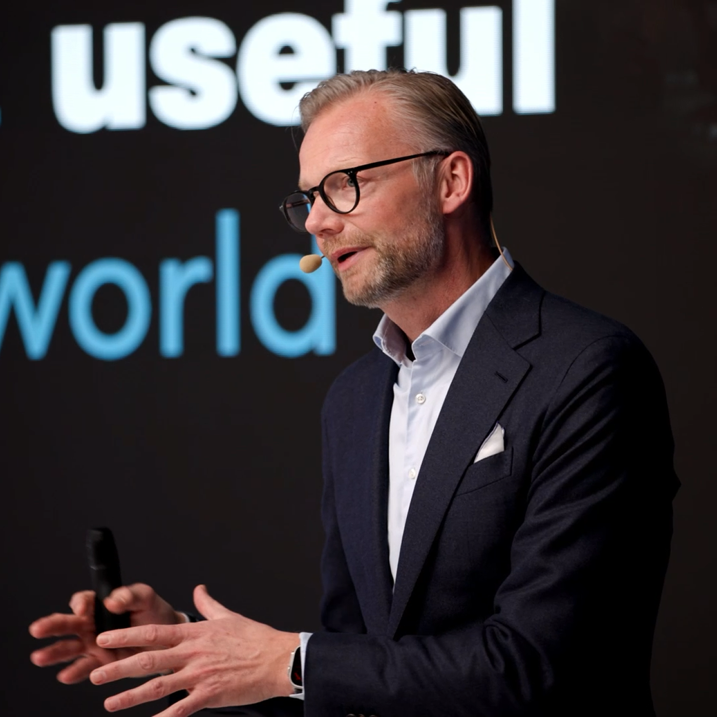 A man in glasses and a suit gestures while speaking on stage at the Trackunit Next event, related to construction IoT.
