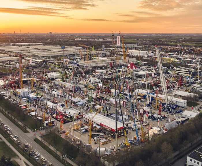 Aerial view of a construction equipment trade show at sunset with cranes, machines, tents, crowds, and fleet management displays.