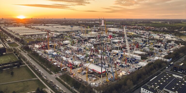 Aerial view of a construction equipment trade show at sunset with cranes, machines, tents, crowds, and fleet management displays.