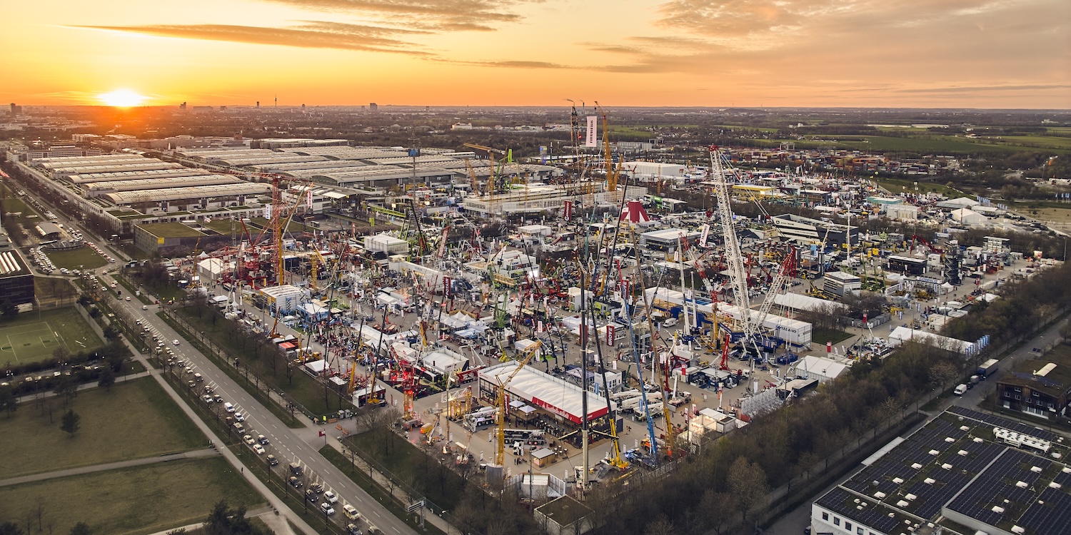Aerial view of a construction equipment trade show at sunset with cranes, machines, tents, crowds, and fleet management displays.