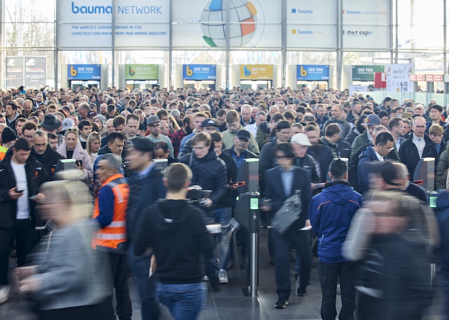 Crowd passing through ticket barriers at a busy trade show entrance, with construction fleet tracking signs in the background.