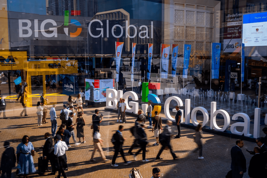 People walk outside a busy event venue with “BIG Global” signage, colorful flags, and glass windows; construction fleet tracking is featured.