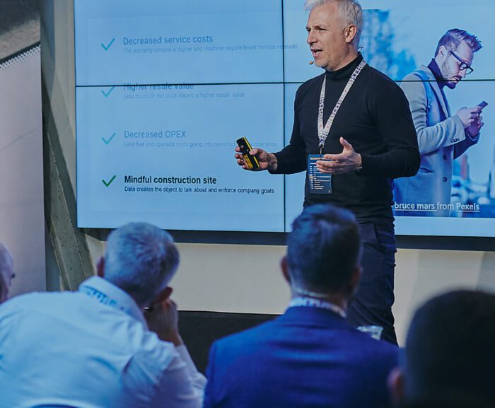 A man presents at the Downtime Summit on service costs and construction, with a screen showing a photo related to connected jobsite.