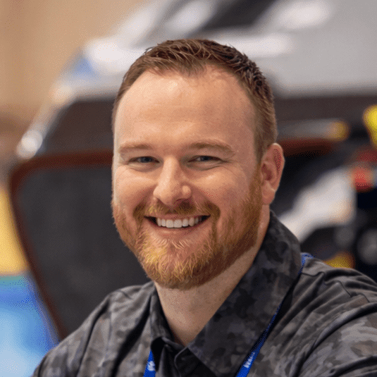 A smiling bearded man in a patterned shirt and Downtime Summit lanyard stands indoors, with a blurred background. Connected jobsite.