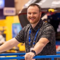 A smiling man with a beard and short hair leans on a blue railing at the Downtime Summit, an indoor connected jobsite event.
