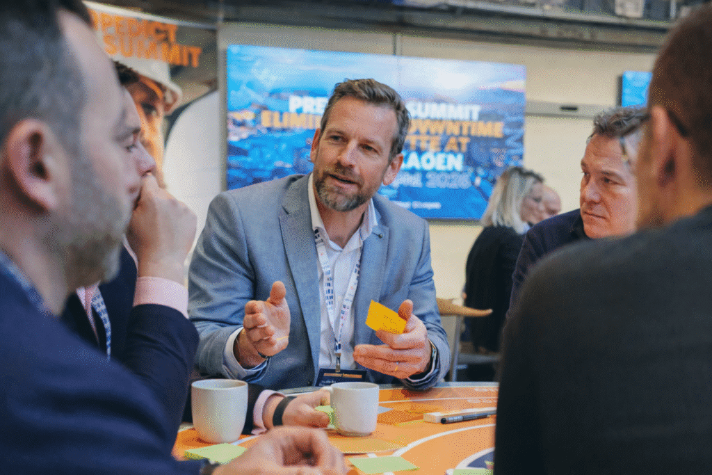 Men in business attire discuss around a table at the Downtime Summit, with coffee, sticky notes, and a presentation screen; connected jobsite.