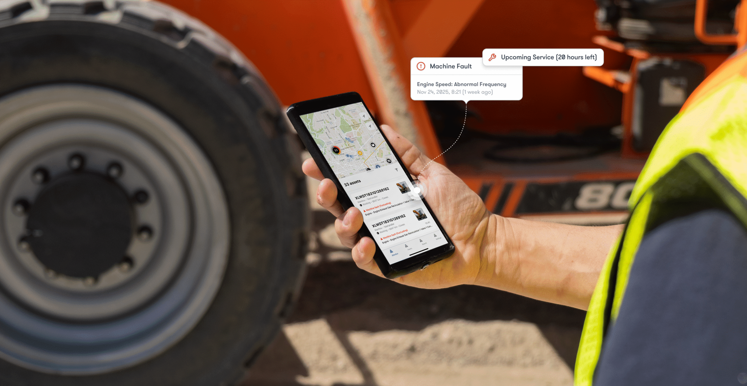 A worker in a yellow safety vest uses a smartphone machine monitoring app with alerts; blurred construction equipment is behind.
