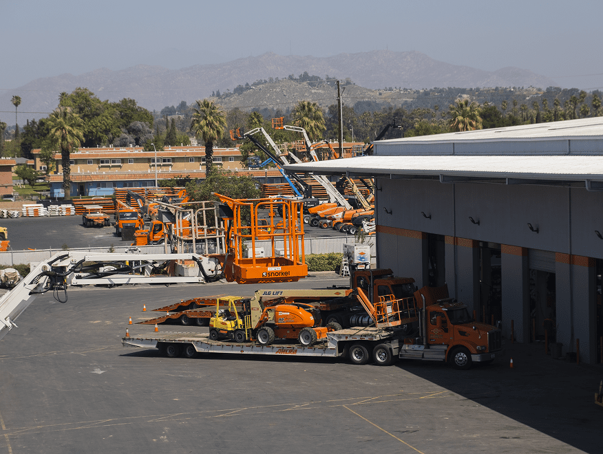 A truck with orange construction equipment is parked by a large industrial building, with more machinery in the lot and mountains behind, showing a connected jobsite.