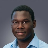 A man with short curly black hair in a light blue shirt smiles at the camera against a gray background at the Downtime Summit, fleet management.