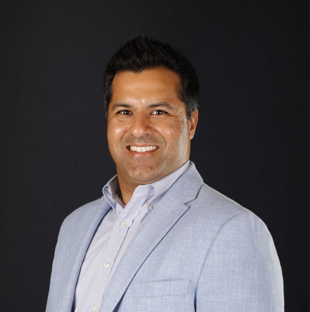A man with short dark hair in a light blue blazer smiles at the camera, ready for the Downtime Summit on construction IoT.