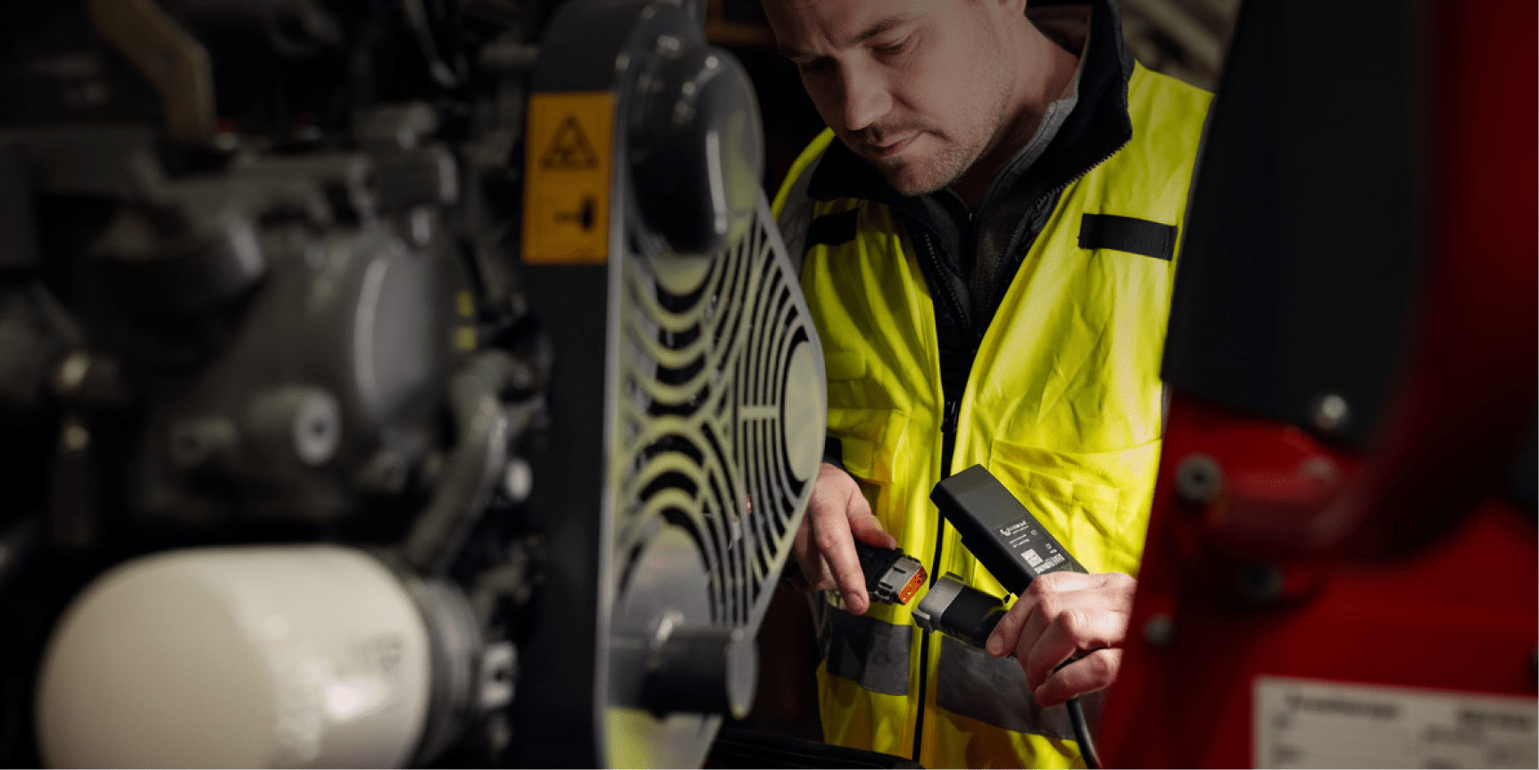 A man in a yellow safety vest inspects machinery with diagnostic equipment at an expo, focused on wires. Construction IoT context.