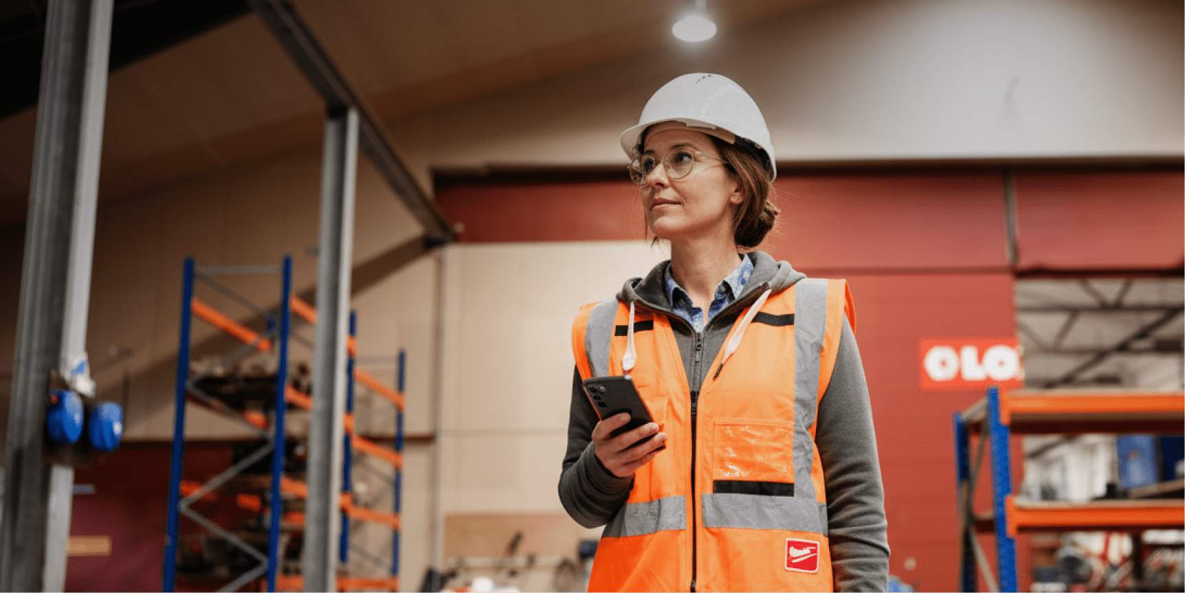 A woman in safety gear oversees installs in a warehouse, using her smartphone on a connected jobsite with industrial shelving behind her.