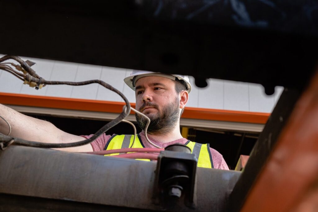 A construction worker in a hard hat and safety vest installs equipment, viewed low-angle with wires in front, showing a connected jobsite.