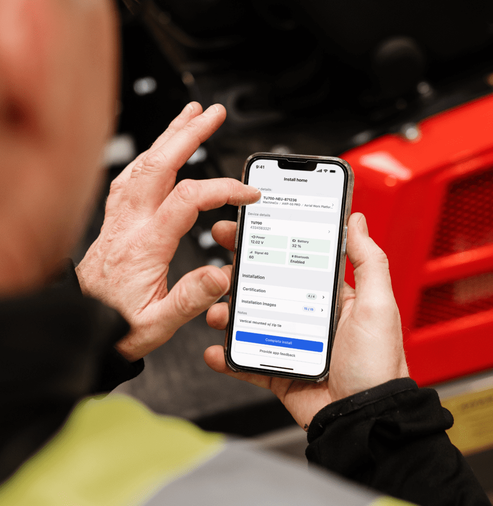 A worker in a yellow safety vest uses a smartphone app near red industrial equipment, illustrating connected jobsite technology.