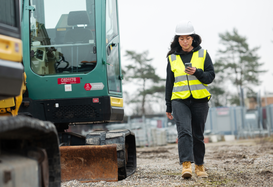 A construction worker in safety gear checks a phone while walking on gravel near a green excavator, illustrating a connected jobsite.