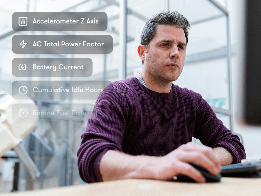 A man in a maroon sweater uses a computer with overlay icons showing telematics metrics for construction equipment tracking.