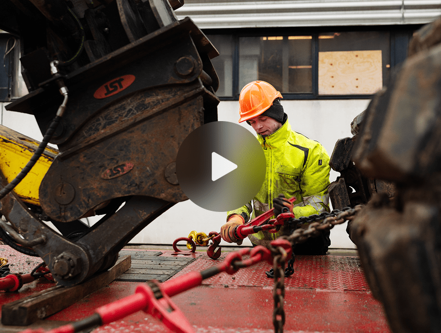 A construction worker secures chains to machinery on a flatbed truck, highlighting construction equipment tracking at a jobsite.