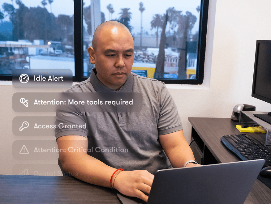 A man in a gray polo works on a laptop with digital alerts onscreen, related to construction equipment tracking; palm trees outside.