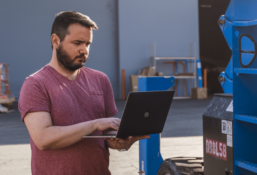 A bearded man in a red t-shirt uses a laptop outdoors, focused, with construction equipment and a warehouse behind him.