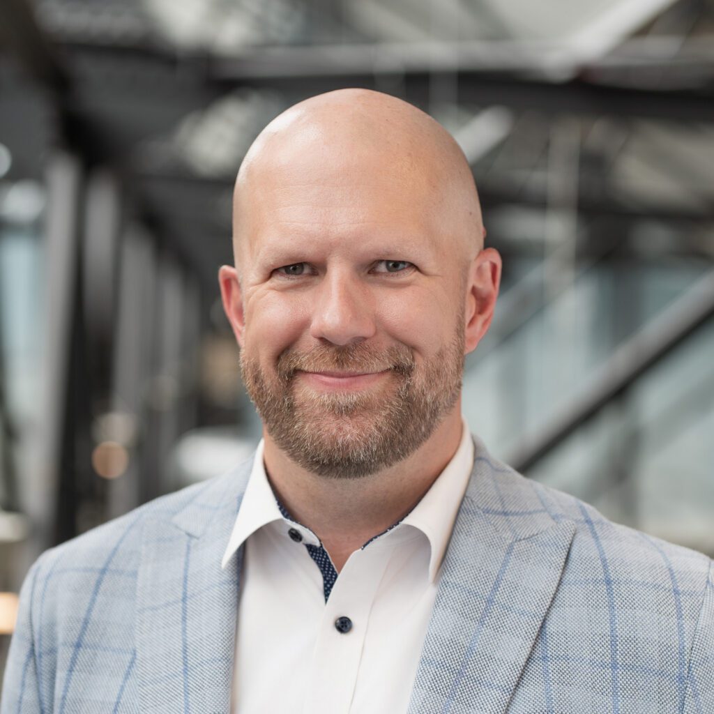 A smiling bald man with a beard, in a gray plaid suit and white shirt, stands in a glass office with the IrisX logo behind him.