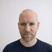 A bald man with a short beard in a dark blue shirt stands against a light background, looking at the camera; construction IoT.