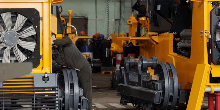 A worker inspects a large yellow machine with construction equipment telematics, surrounded by heavy machinery in a busy factory.