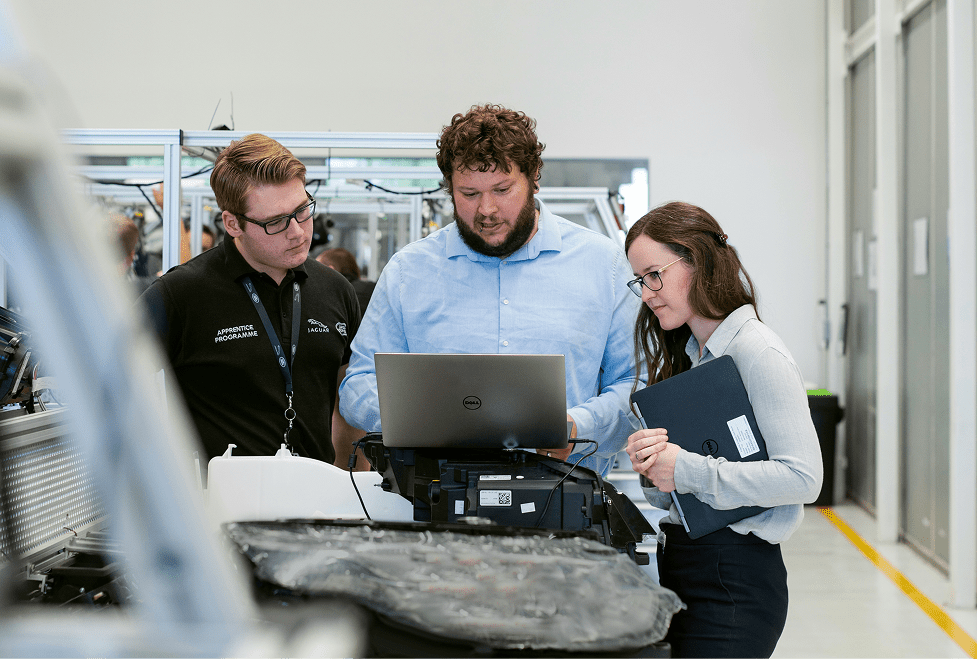 Three people in an industrial setting discuss work while looking at a laptop, surrounded by equipment for construction telematics.
