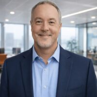 A man with short gray hair and a goatee, in a blue suit, smiles in a modern office, suggesting a connected jobsite setting.