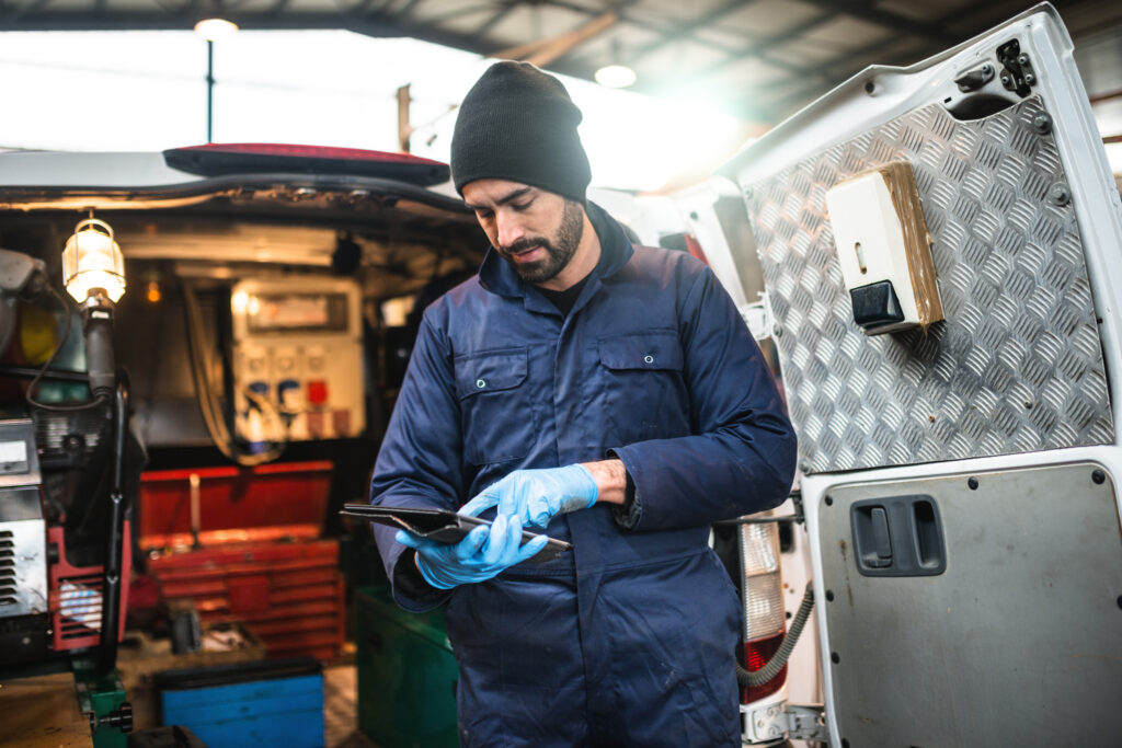 A mechanic in blue uses a tablet by an open van in a workshop, with tools inside and around, highlighting fleet management.