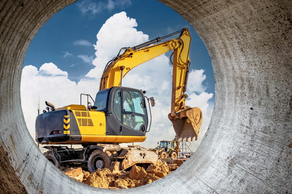 A yellow excavator digs on a construction site, seen through a concrete pipe, with another vehicle and blue sky—connected jobsite.