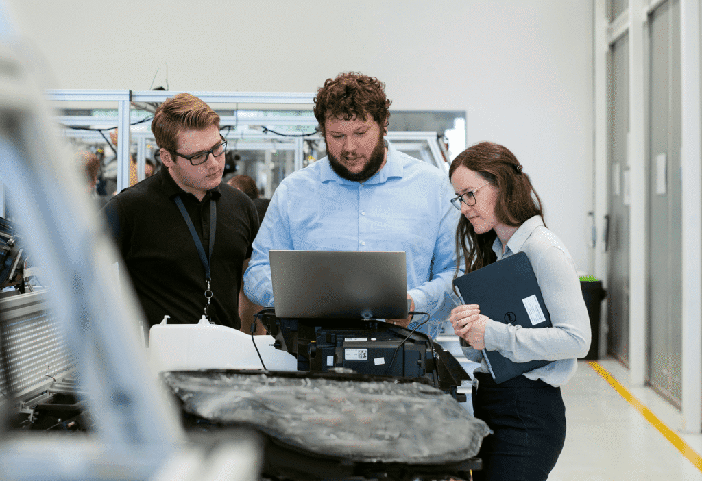 Three people in a bright industrial workspace gather around a laptop on equipment, focused on construction equipment tracking.