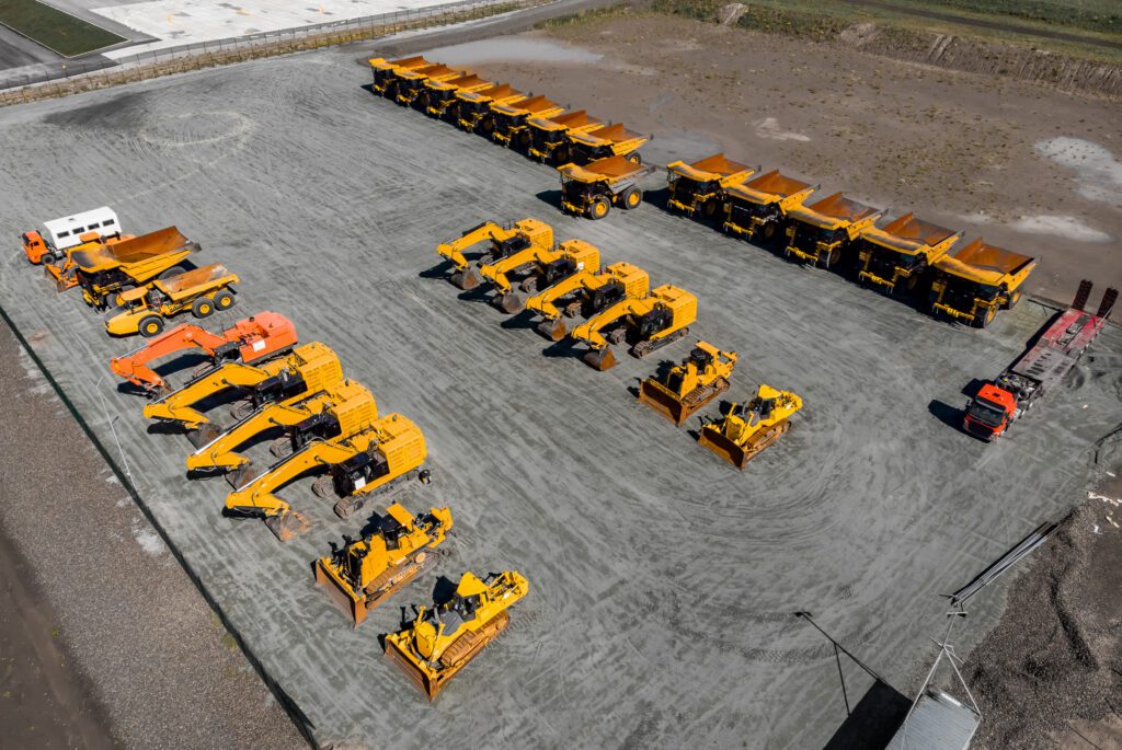 Aerial view of a construction equipment fleet neatly arranged on a paved lot, with two orange trucks at opposite corners.