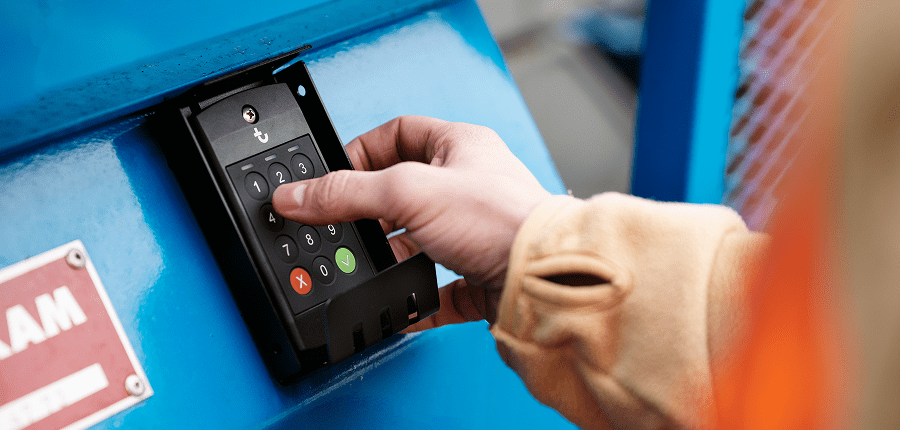 A person in a beige sleeve and orange safety vest uses a black keypad on a blue metal surface, showing connected jobsite access.