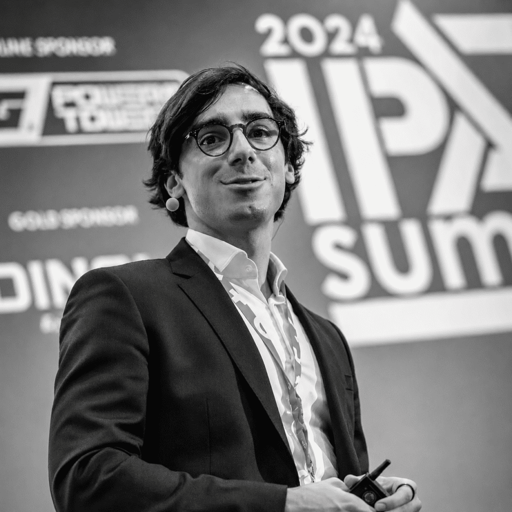 A man in a suit and glasses with a headset speaks on stage at the 2024 Downtime Summit, related to construction IoT. Black and white.