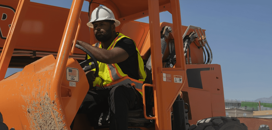 A construction worker in a white hard hat and yellow vest operates an orange forklift outside, illustrating connected jobsite.