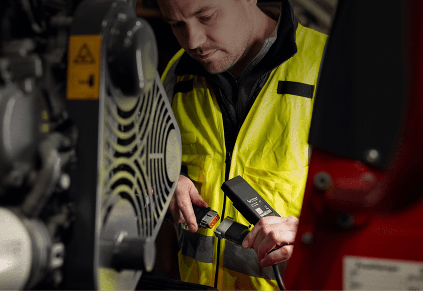 A man in a yellow safety vest inspects industrial machinery with a handheld device, illustrating construction equipment tracking.