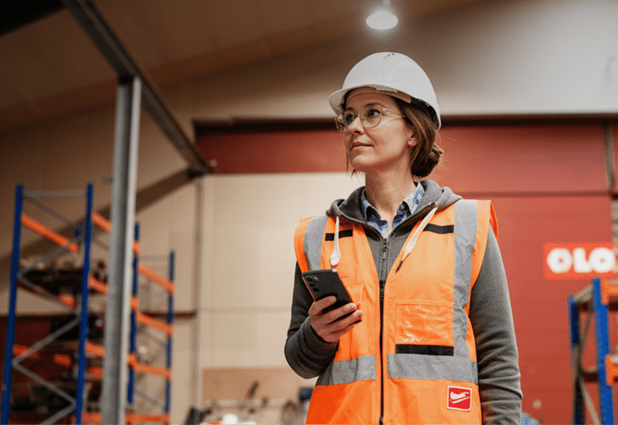 A woman in safety gear with a smartphone stands indoors near industrial shelves, suggesting a connected jobsite environment.