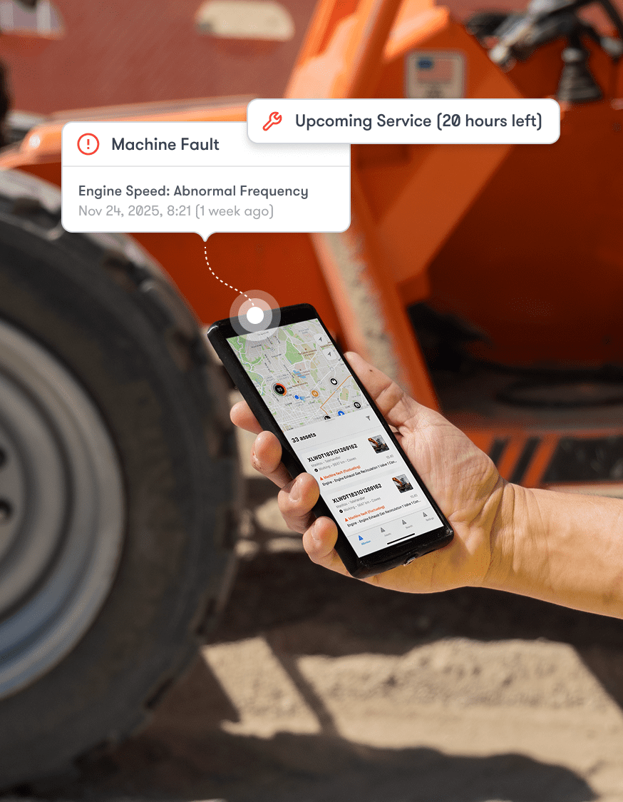 A person holds a smartphone with machine alerts and map; a large orange equipment tire is in the background, showing construction IoT.
