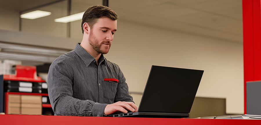 Bearded man in a striped shirt types on a black laptop at a red counter in a modern office, related to construction equipment tracking.