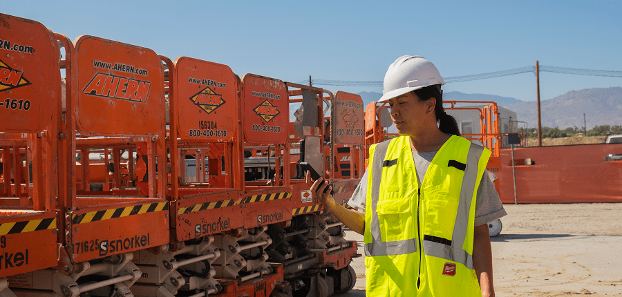 A construction worker in a hard hat and safety vest uses a smartphone by orange scissor lifts at a sunny connected jobsite.