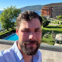 A man with short brown hair and a beard takes a selfie outdoors near a sunny lakeside resort, pool, and green lawn.