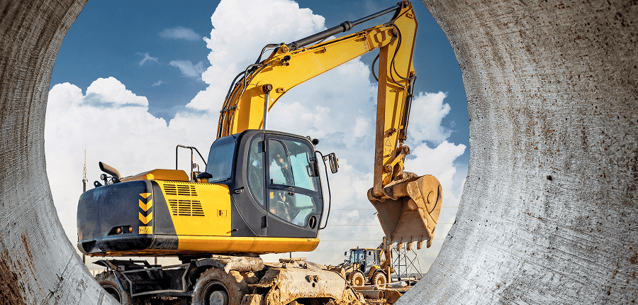 A yellow excavator on a construction site seen through a large pipe, with another vehicle in the distance and blue sky overhead.