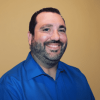 A smiling man with short dark hair and a beard in a blue collared shirt, pictured against a light brown background, related to fleet management.