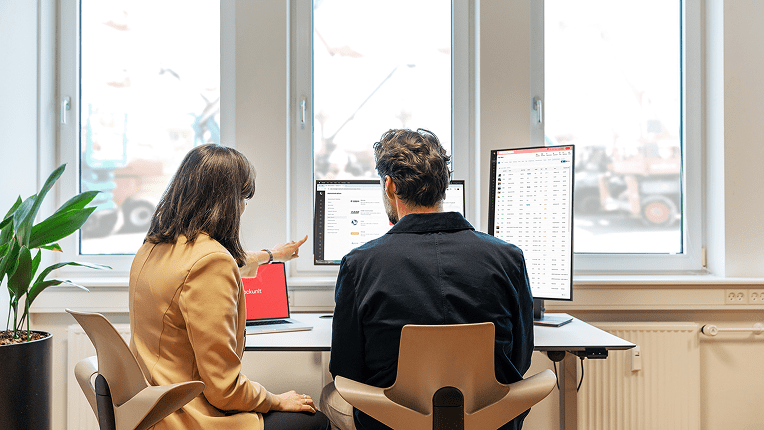 Two people at a desk review data on laptops and monitors in a customer portal, discussing construction equipment tracking.
