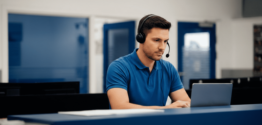 A man in a headset and blue polo shirt works on his laptop at a desk in a modern office, suggesting a connected jobsite setting.