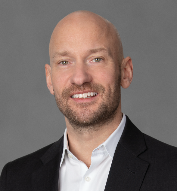 A bald man with a trimmed beard, in a dark suit and white shirt, smiling before a gray background, representing construction telematics.