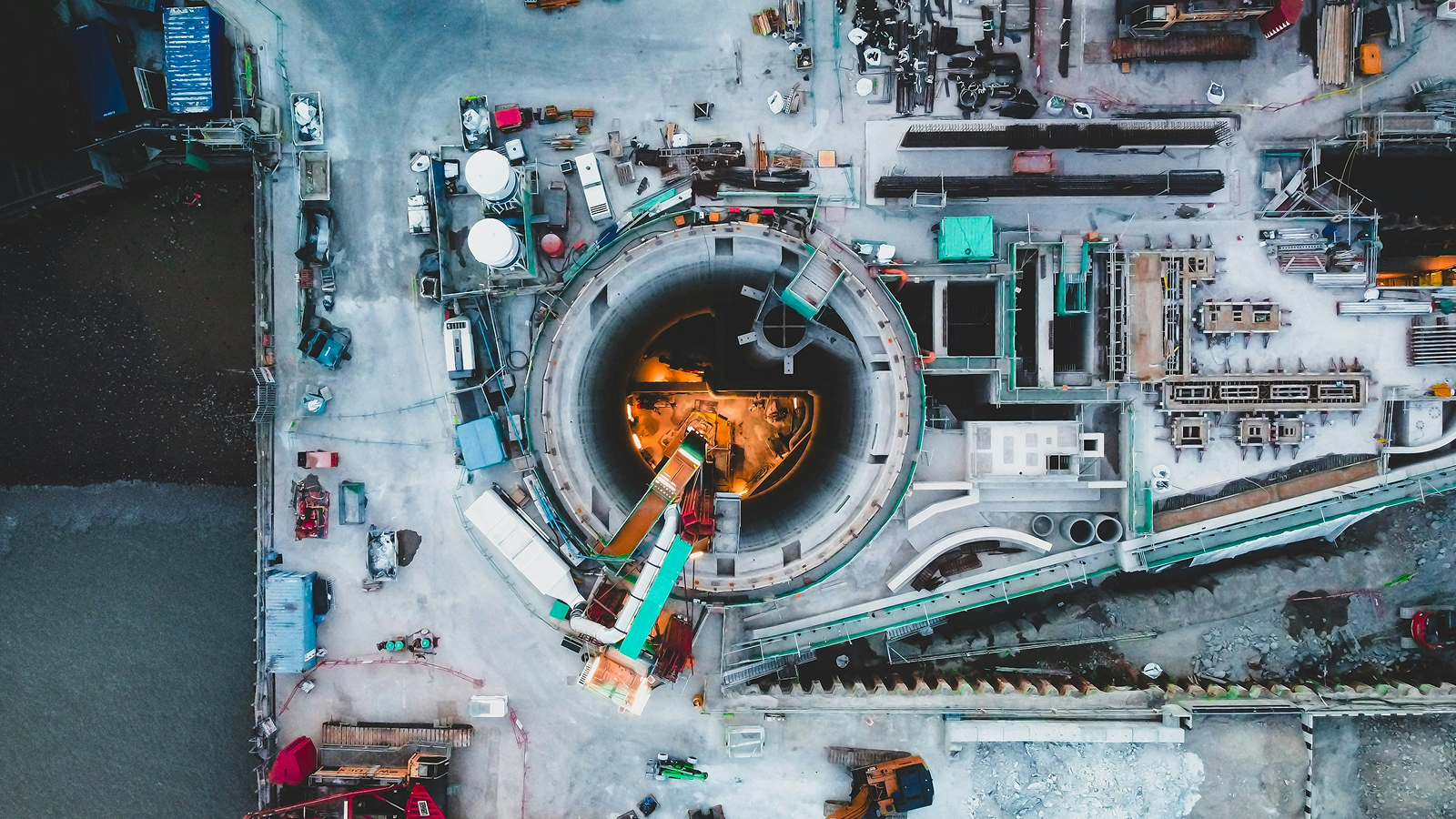 Aerial view of a construction site with a circular tunnel entrance, machinery, and workers; bright lights highlight a connected jobsite.